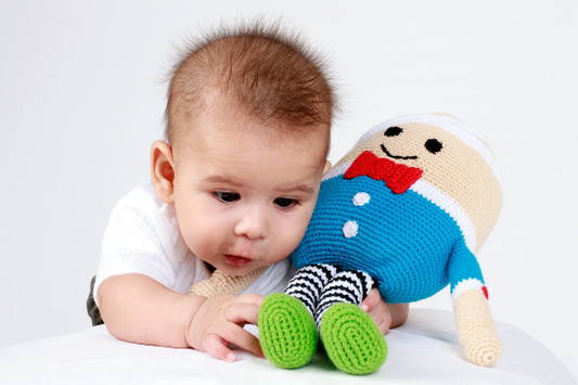 A baby is playing with a crochet doll with a striped body, blue shirt, and red bow tie, with a bandaged head.