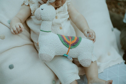 A baby holding a crochet toy resembling a cream-colored horse with purple and orange details