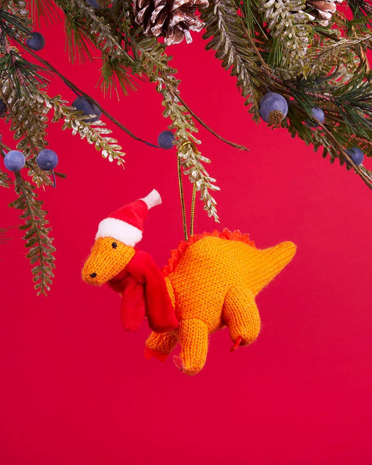 Knitted dinosaur ornament with a Santa hat hanging from Christmas tree branches on a red background