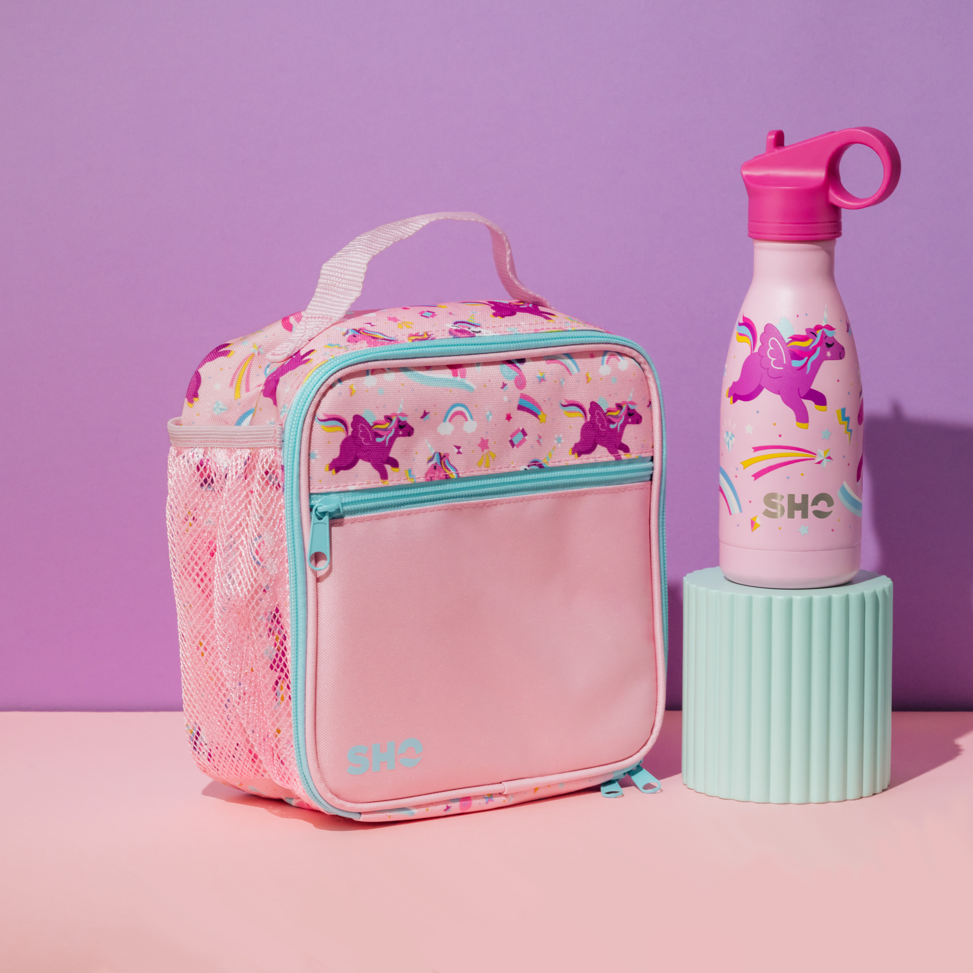 Pink lunch bag with unicorn design and pink water bottle with 'SHO' branding on a purple background