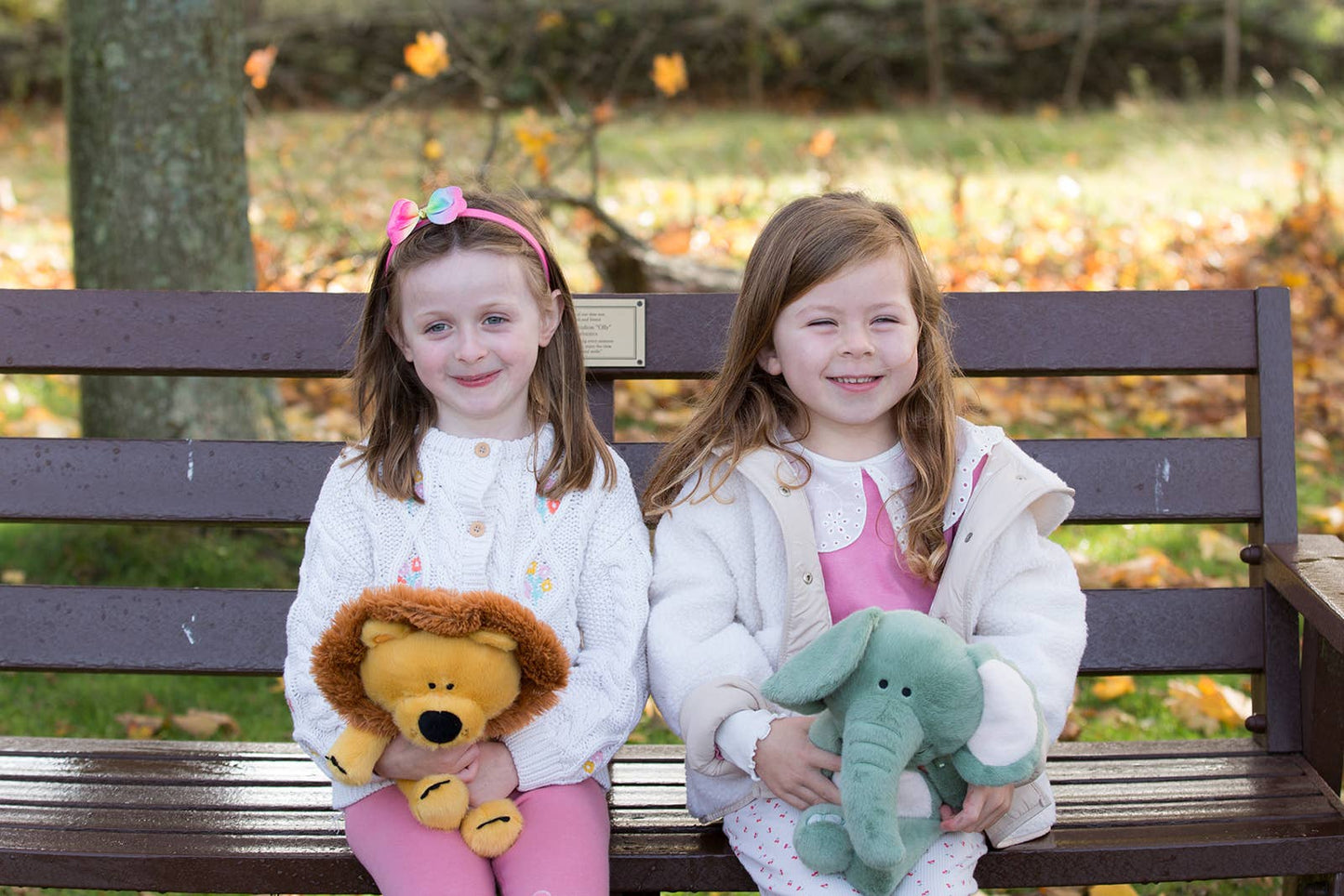 Two young girls sitting on a bench with stuffed animals in an outdoor setting.