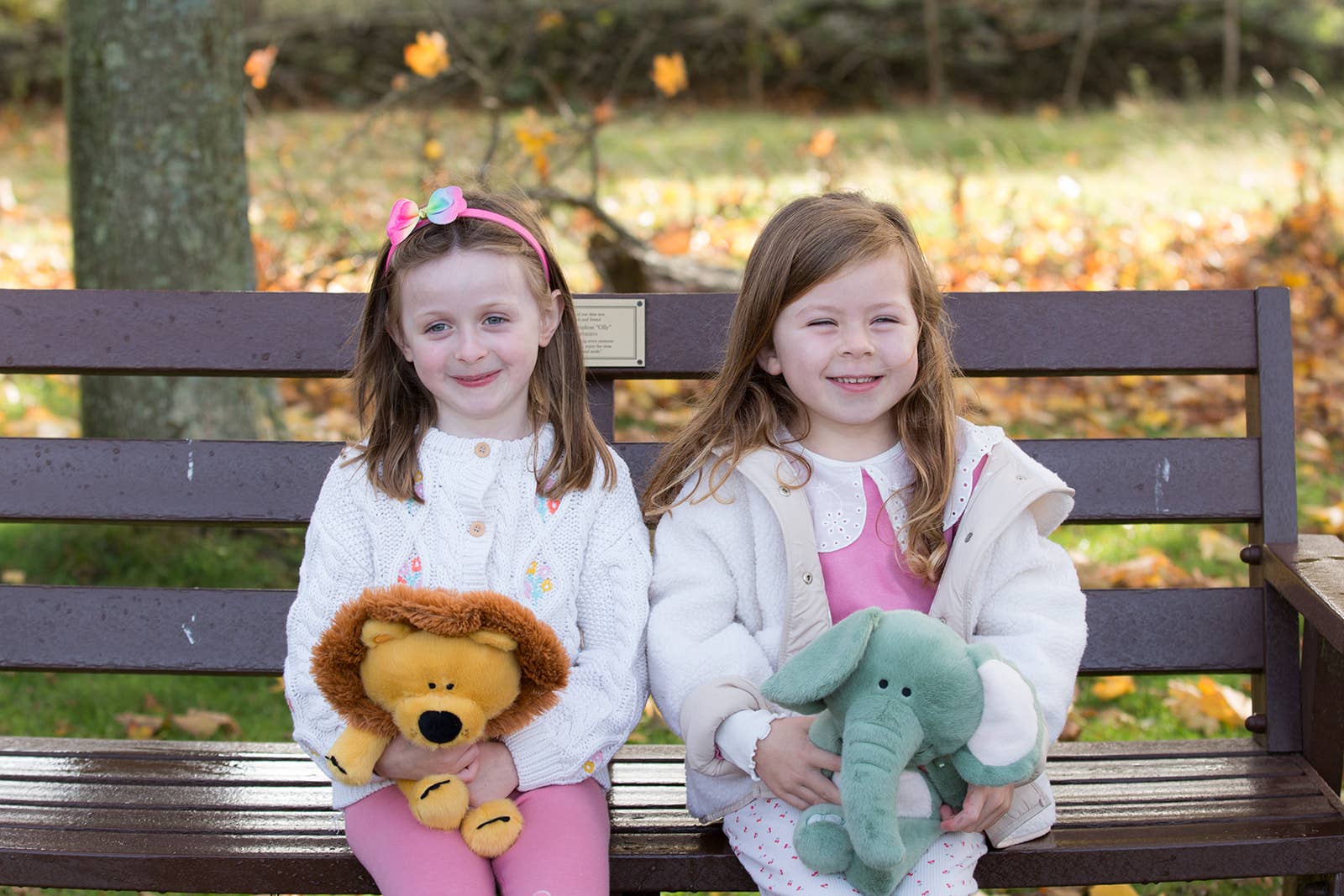 Two young girls sitting on a bench with stuffed animals in an outdoor setting.