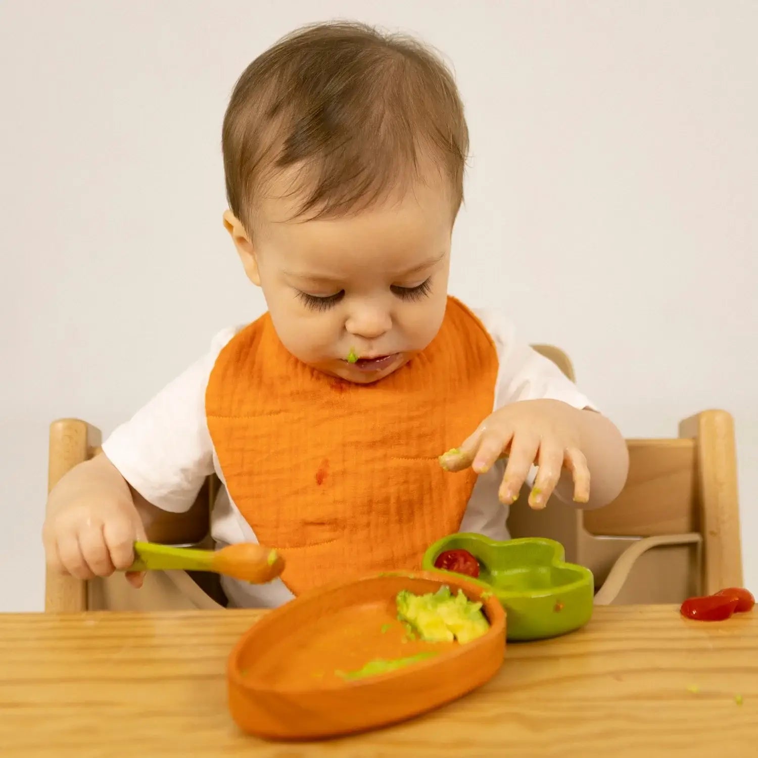 Baby in an orange bib eating with a wooden high chair and colorful plates.
