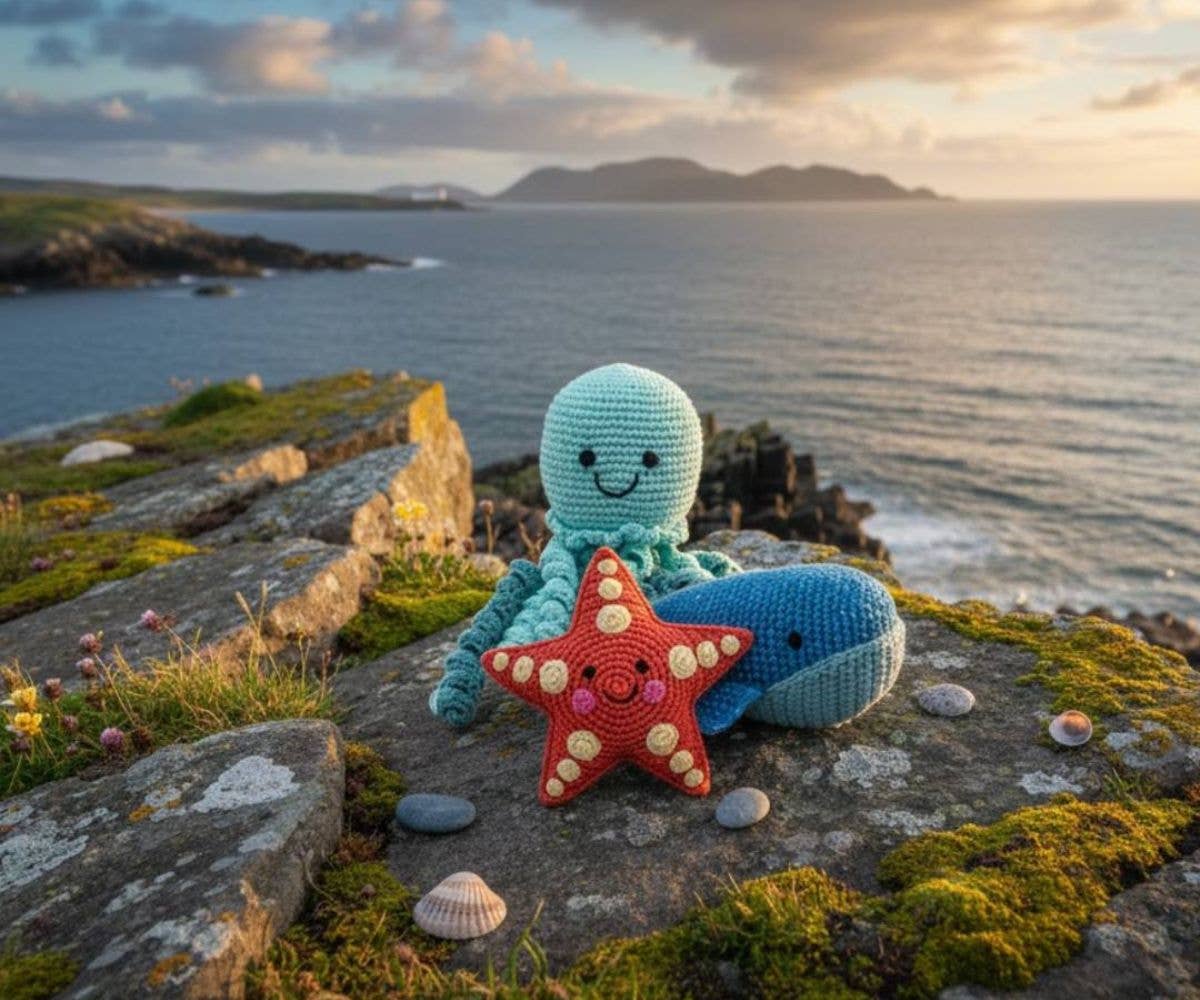 Crocheted octopus, whale and starfish toy sitting on a rocky coastal landscape with ocean and sunset.