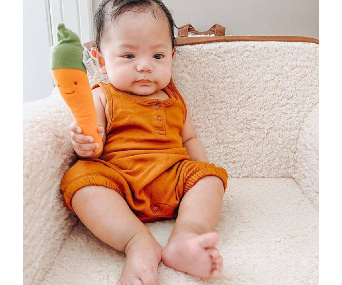 Baby in orange outfit holding a carrot-shaped toy on a white surface