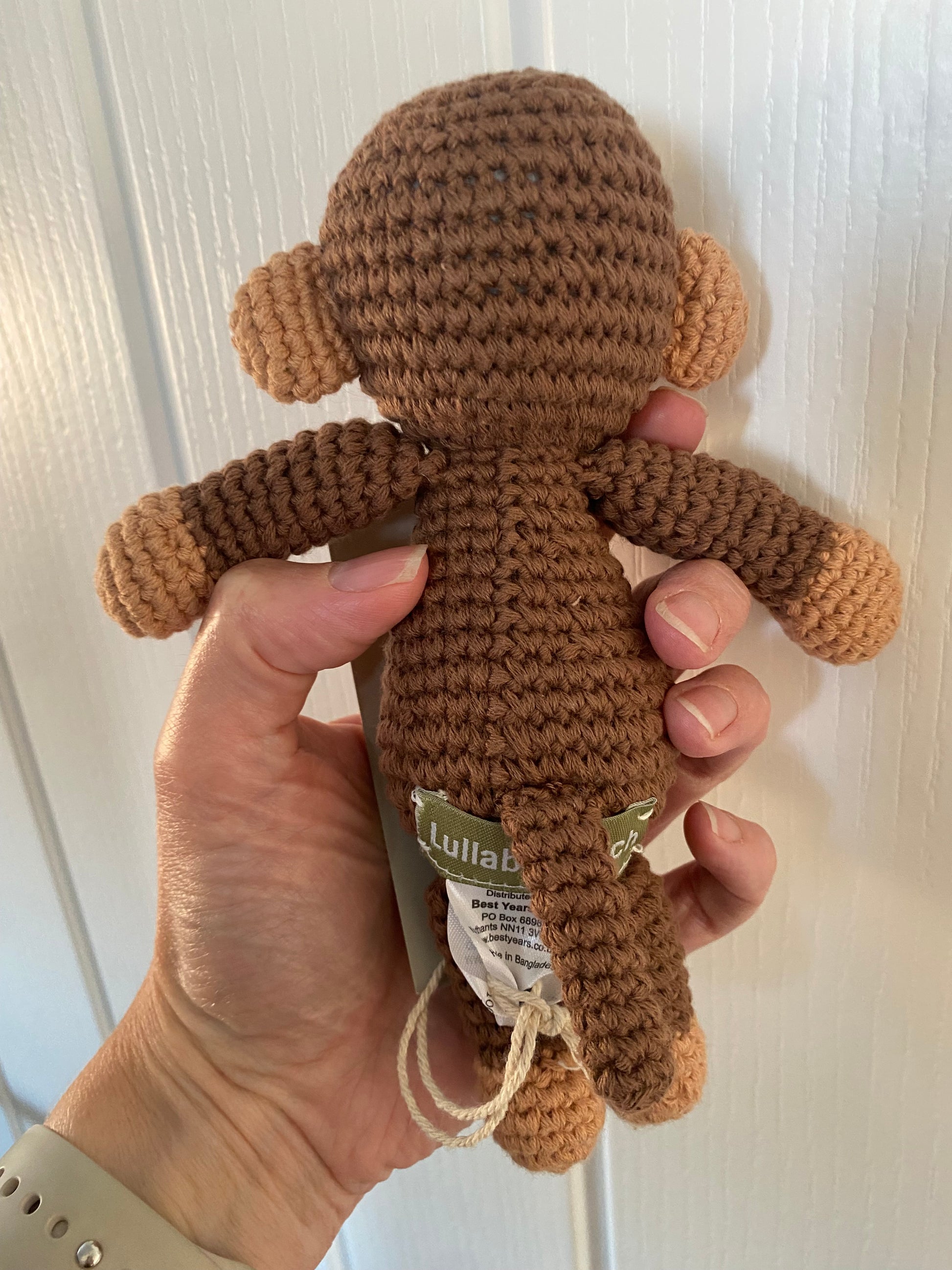 A hand holds a small crocheted monkey toy against a white paneled door. The toy is primarily medium brown with lighter tan accents on the ears, hands, and feet.