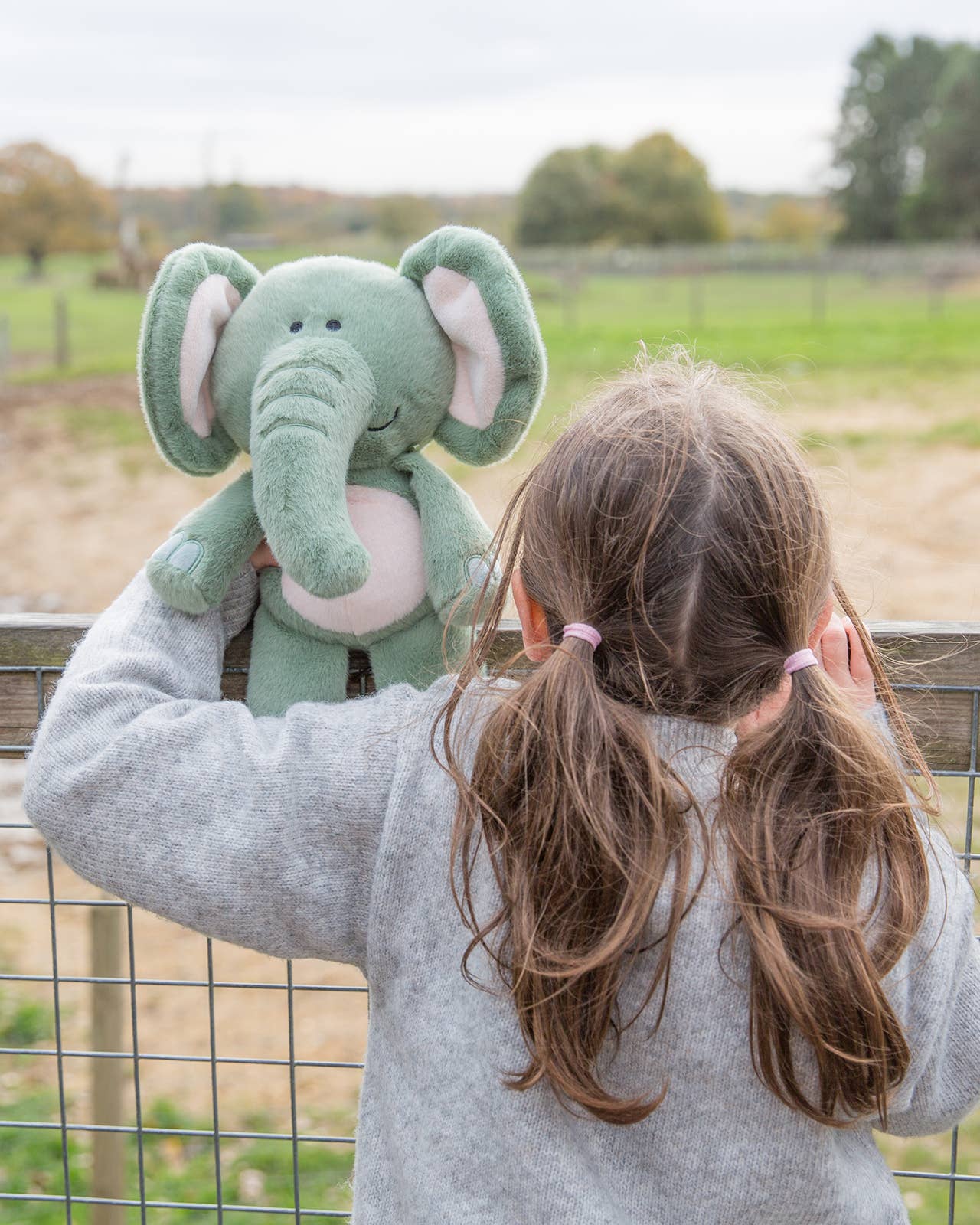 Child holding a large green stuffed elephant outdoors with a fence and trees in the background