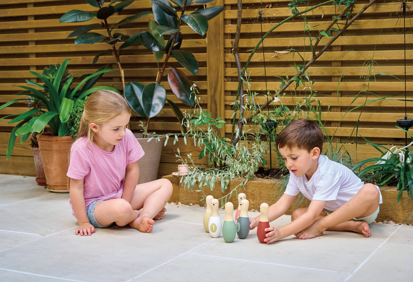 Two children playing with colorful wooden toys on a patio surrounded by plants.