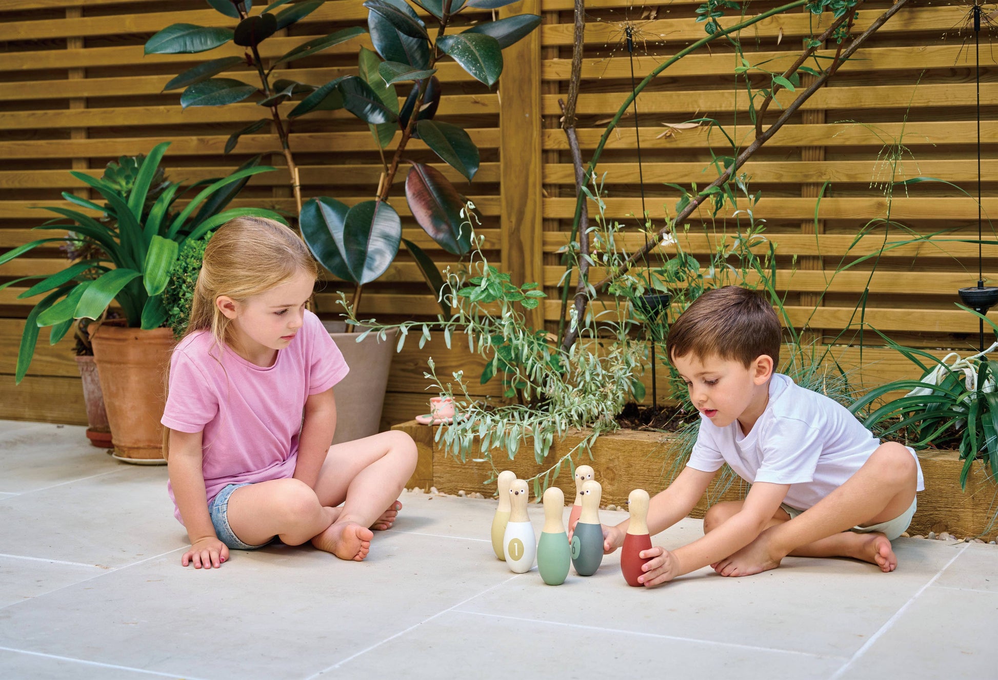Two children playing with colorful wooden toys on a patio surrounded by plants.