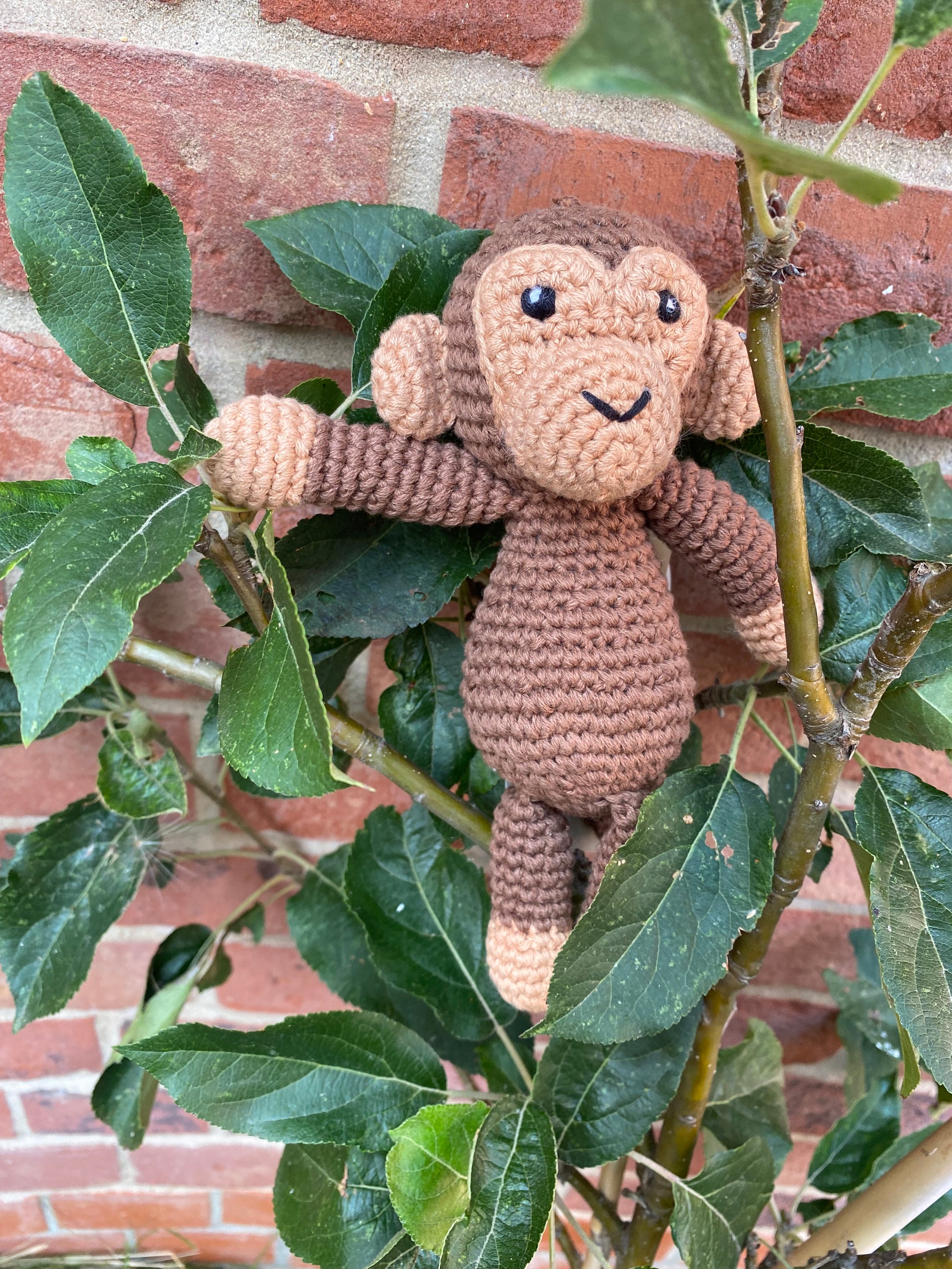 A crocheted plush monkey toy in shades of brown and tan, hanging among green leaves in front of a red brick wall.