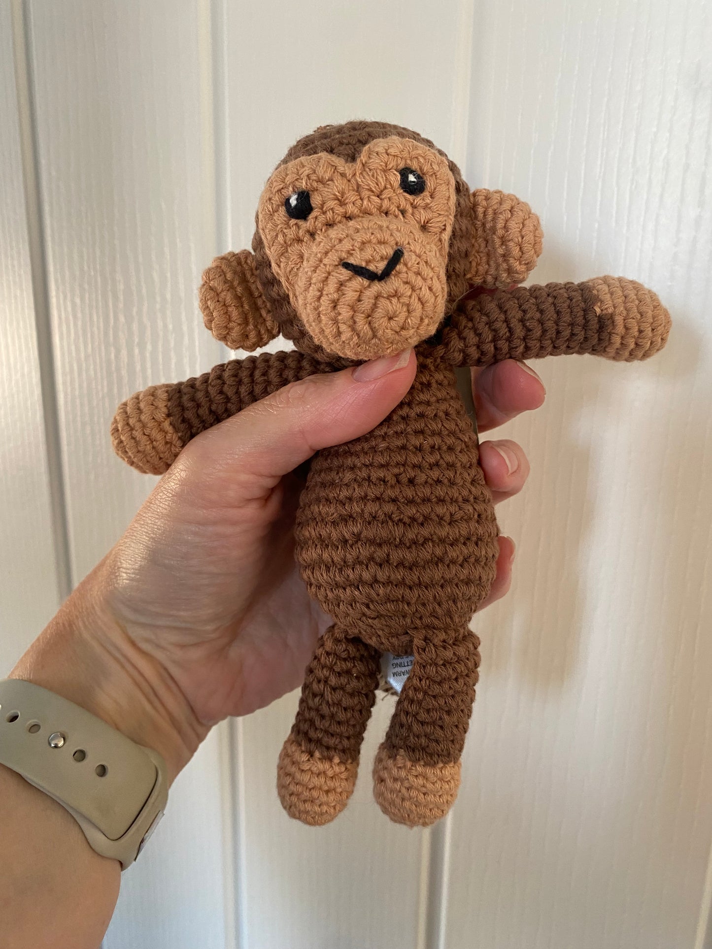 A hand holds a small crocheted plush monkey toy against a white paneled door background. The toy is brown with lighter tan accents on its face, hands, and feet, and has black embroidered eyes and a stitched smile.