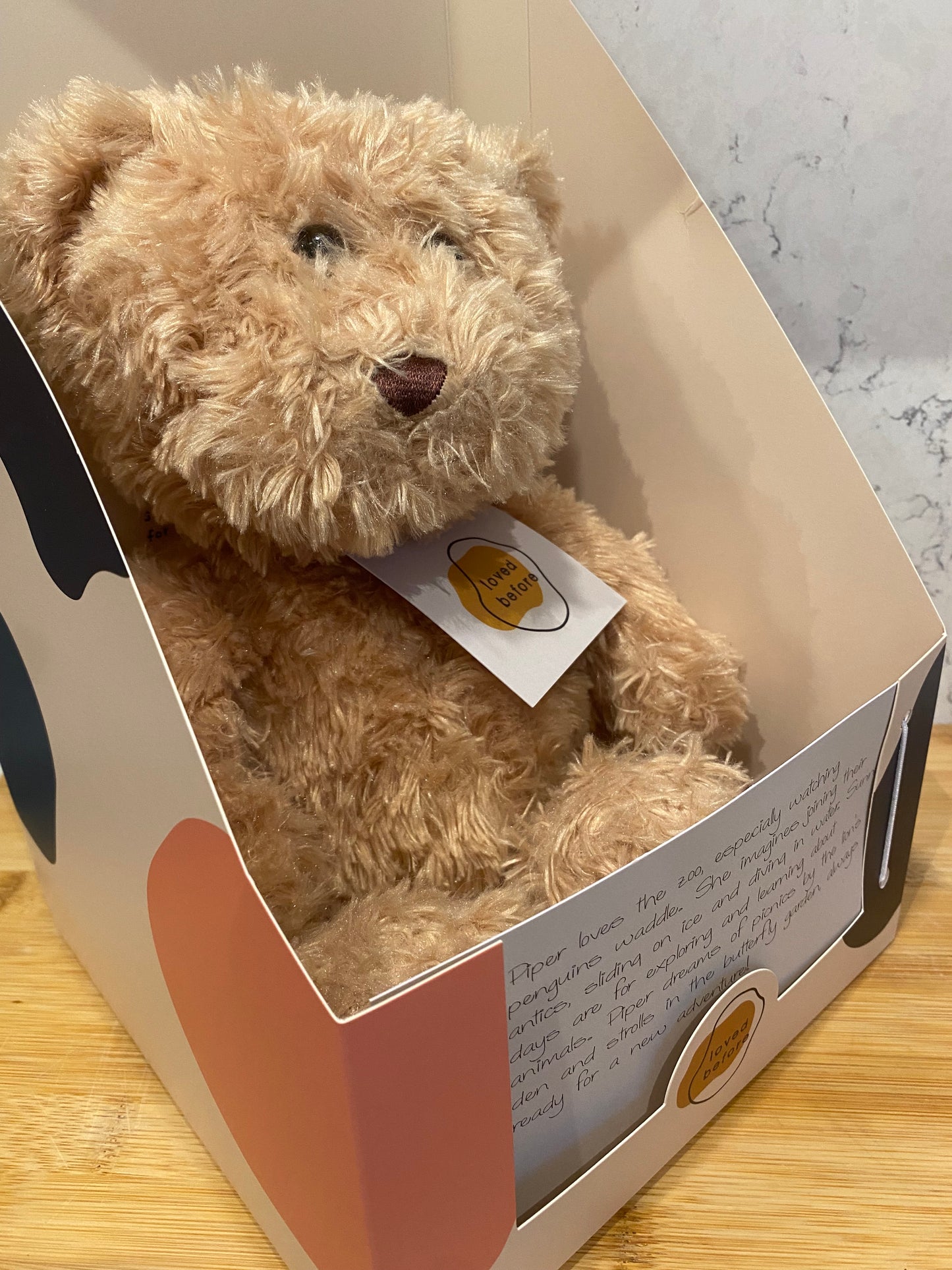 Brown teddy bear inside an open box with a visible brand label on a wooden surface.