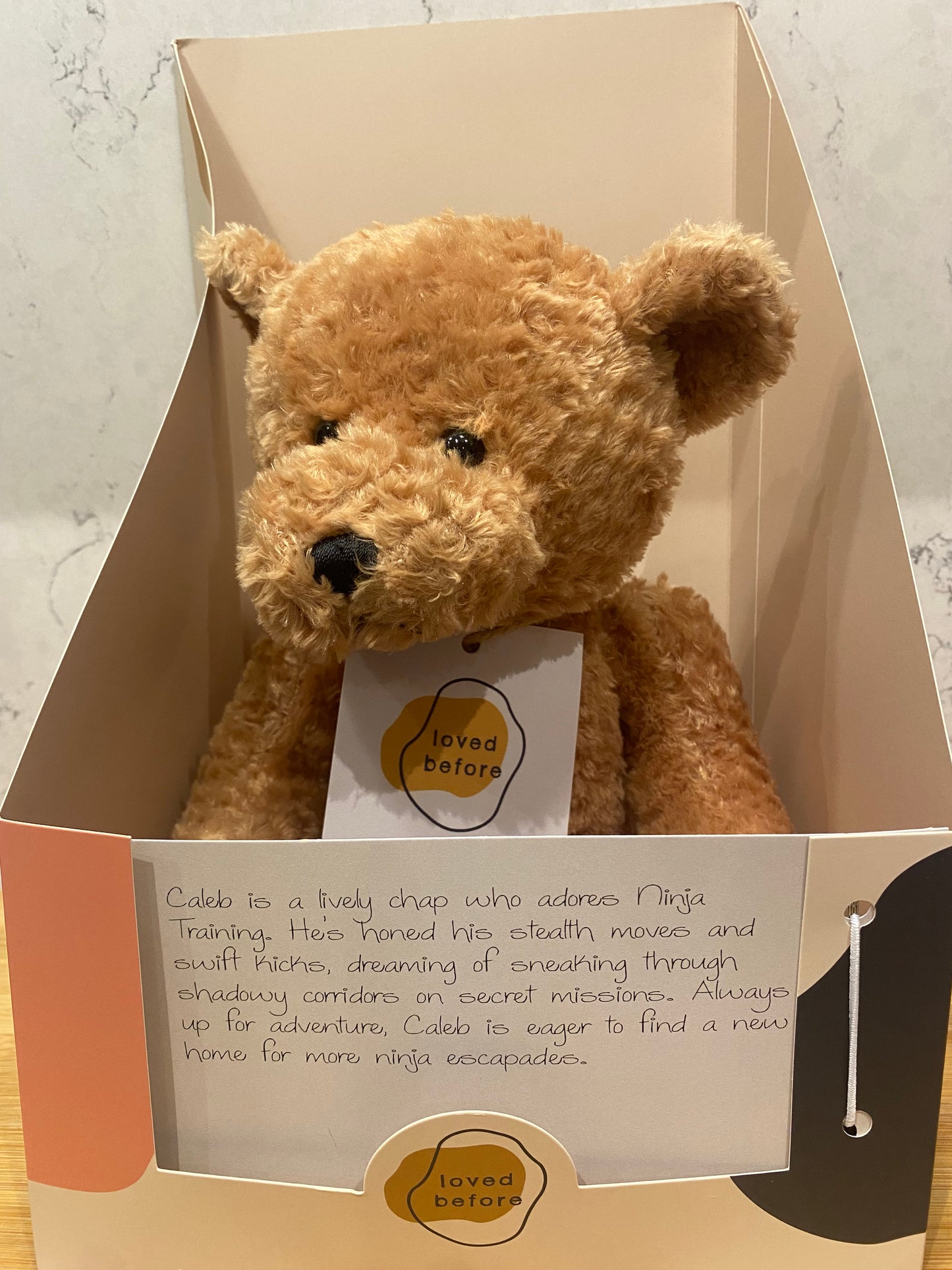 Brown teddy bear in a box with a label on a light-colored surface