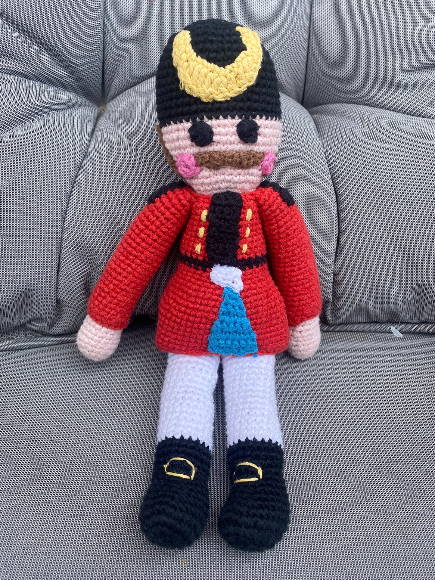 Crocheted toy figure of a character with a red outfit and black shoes on a gray couch.