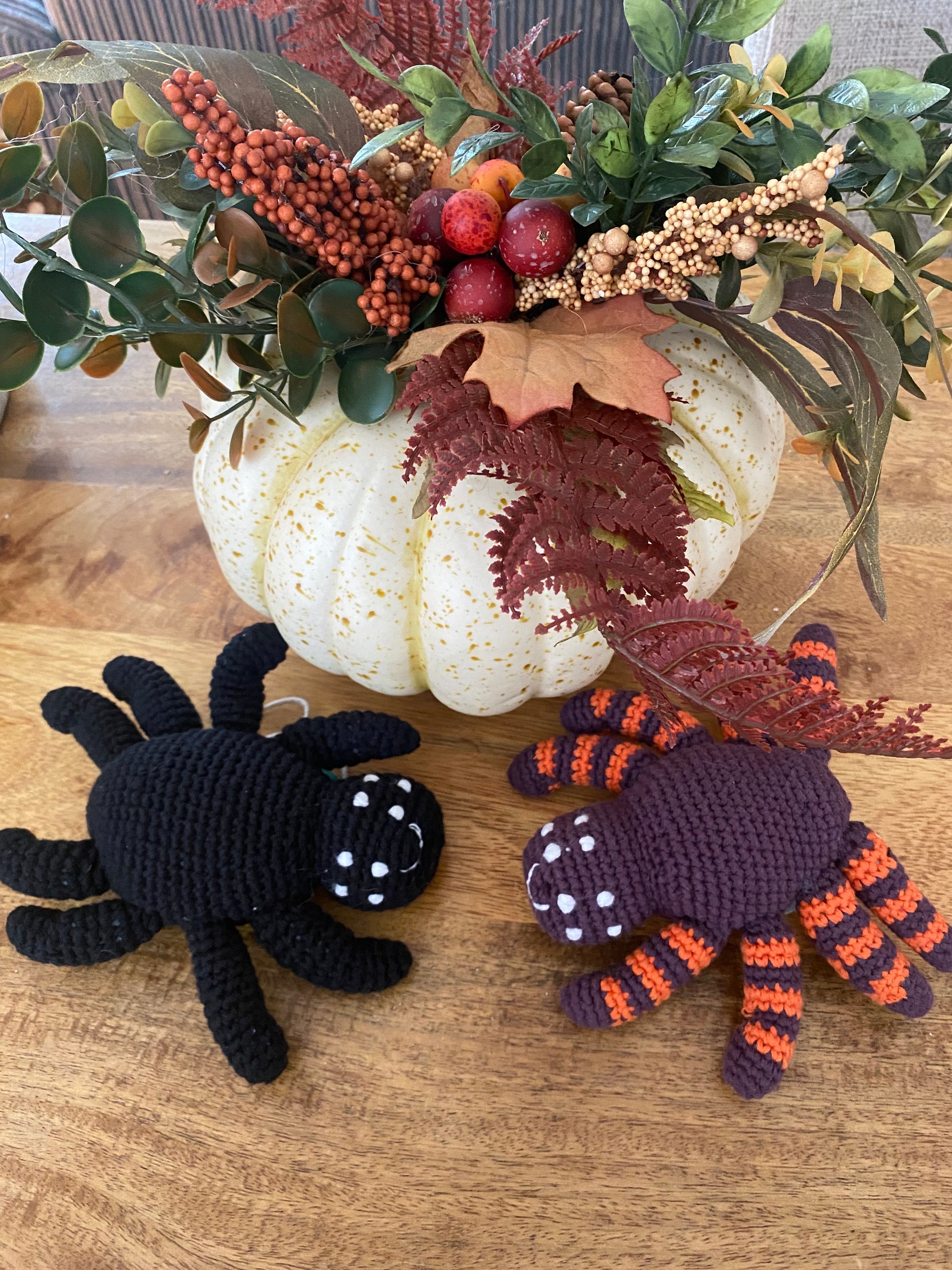 Two crocheted spiders on a wooden surface with pumpkins and foliage.