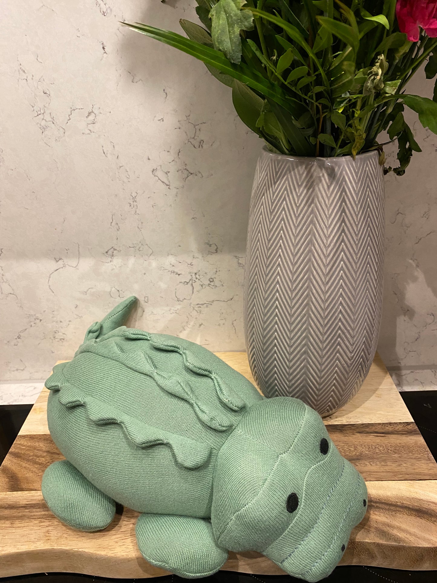 A large plush toy resembling a green crocodile made from organic cotton, suitable for babies, next to a grey vase of flowers.