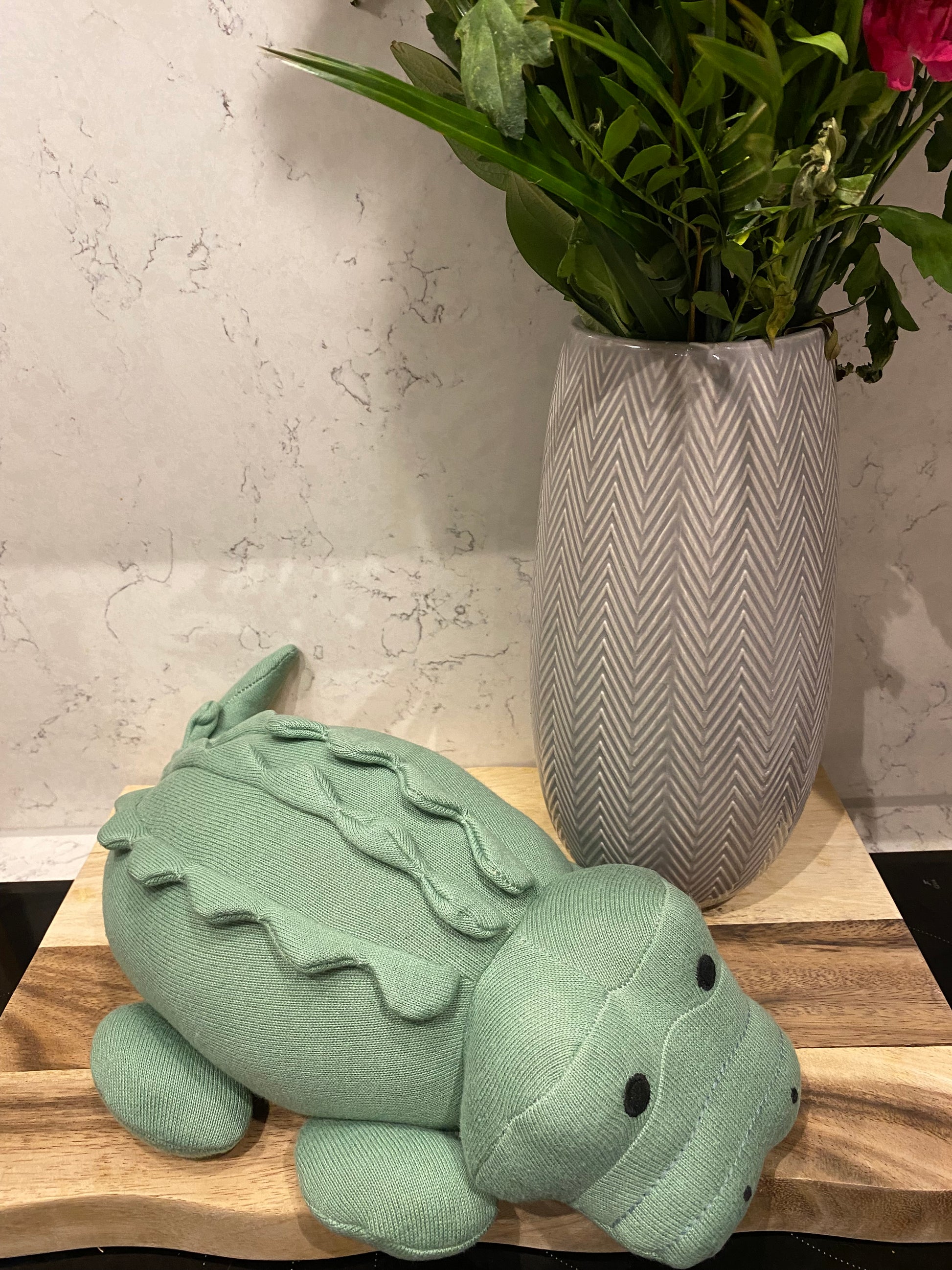 A large plush toy resembling a green crocodile made from organic cotton, suitable for babies, next to a grey vase of flowers.
