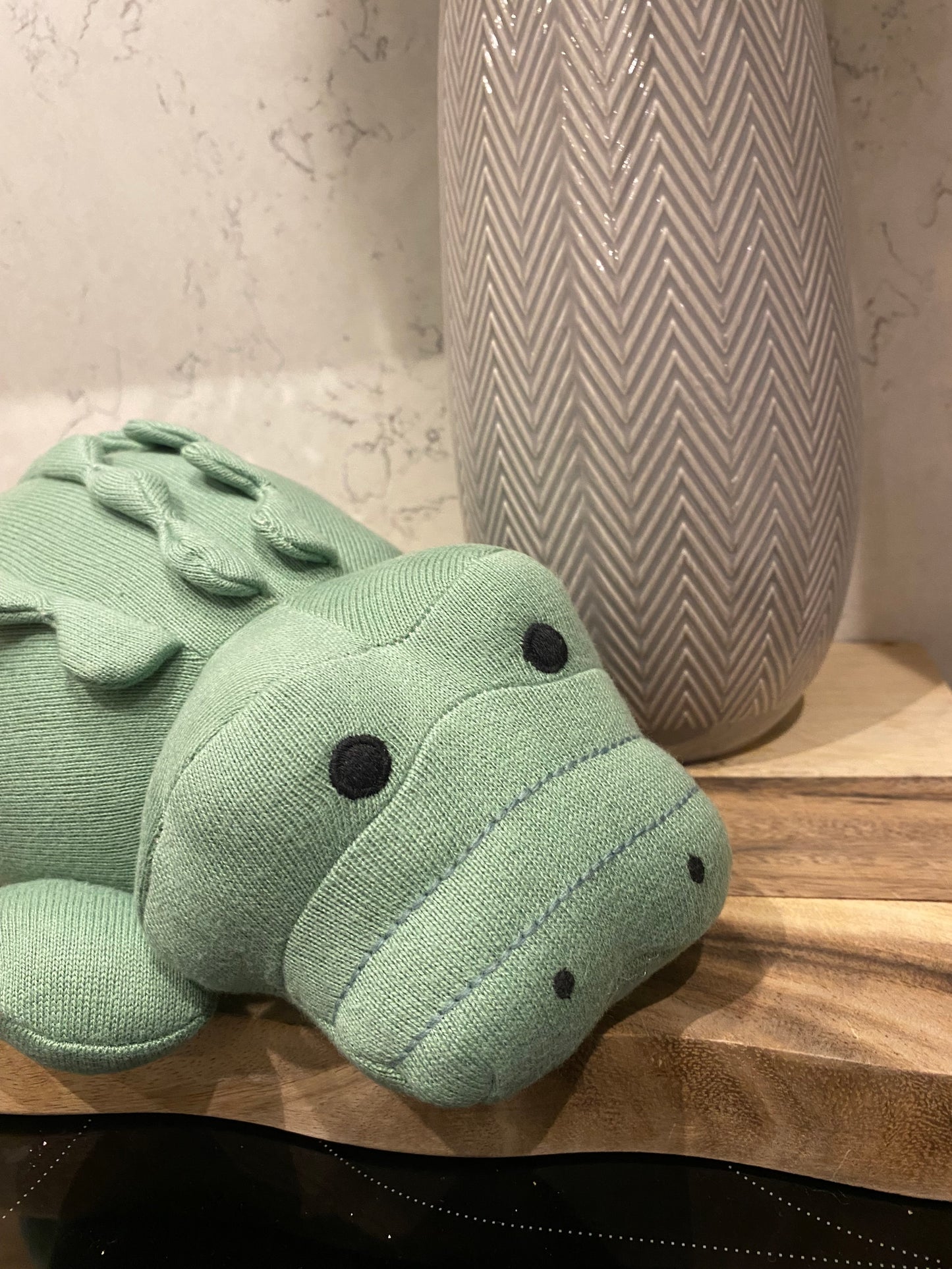 A large plush toy resembling a green crocodile made from organic cotton, suitable for babies, next to a grey vase of flowers
