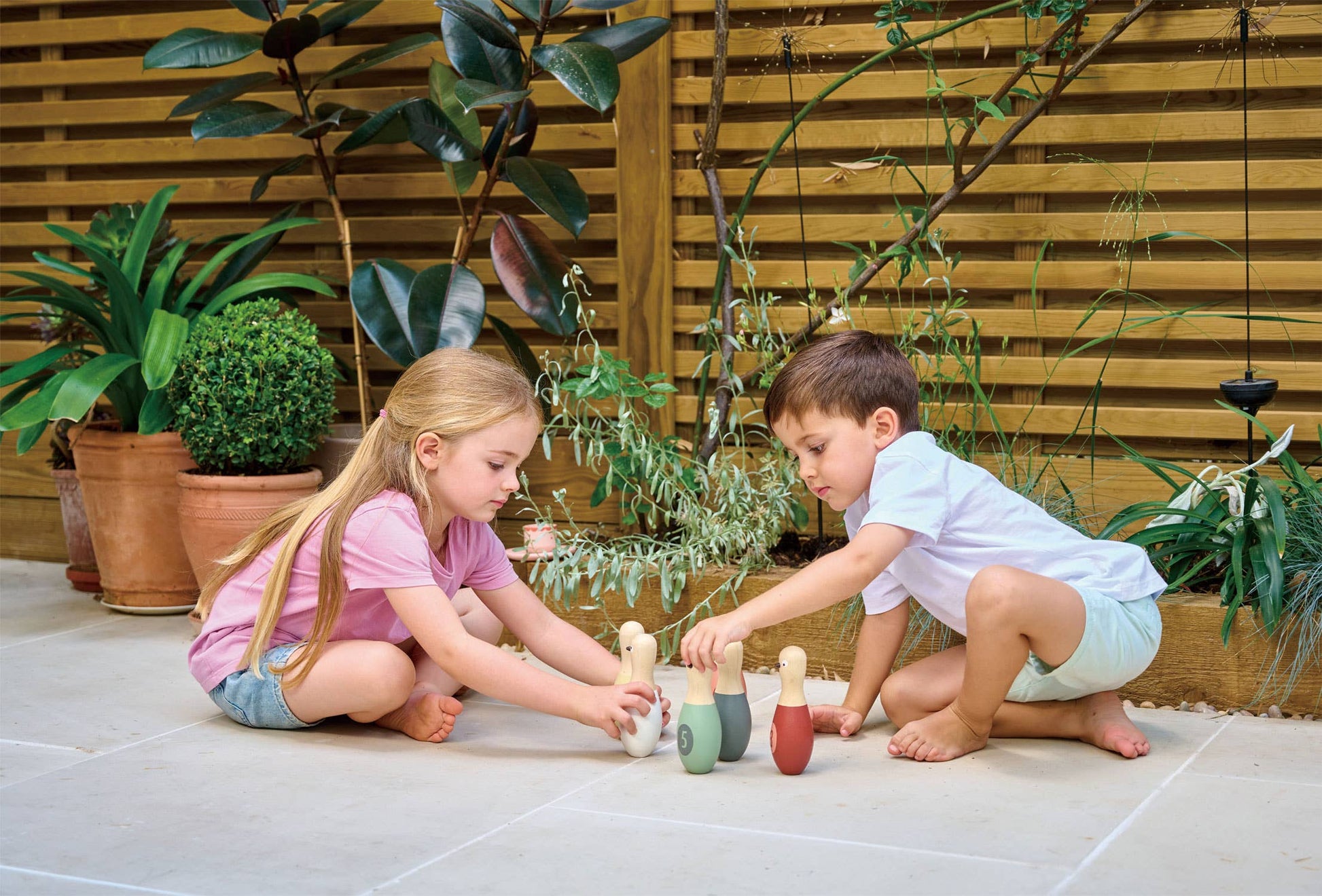 Two children playing with wooden toys outdoors on a stone patio surrounded by plants.