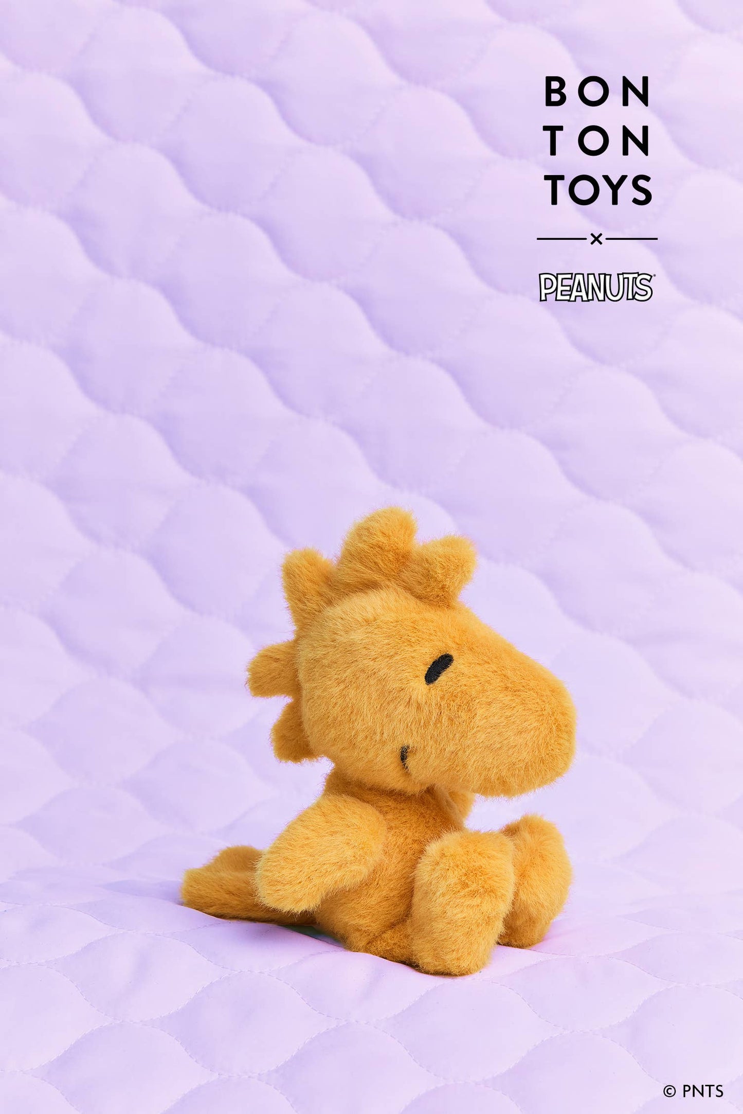 A small yellow plush toy resembling Woodstock sits on a lavender quilted surface. The image includes the text 'BONTON TOYS x PEANUTS' in the upper right corner.