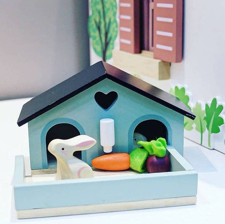 Toy rabbit with vegetables and a bottle inside a small house on a white surface.
