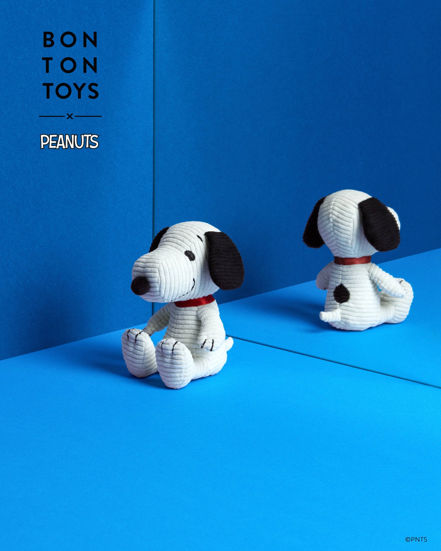 A crocheted plush toy of Snoopy from Bon Ton Toys x Peanuts, shown in a studio setting with its reflection visible in a blue mirror. The background is a solid blue with the 'BON TON TOYS x PEANUTS' logo in the upper left.