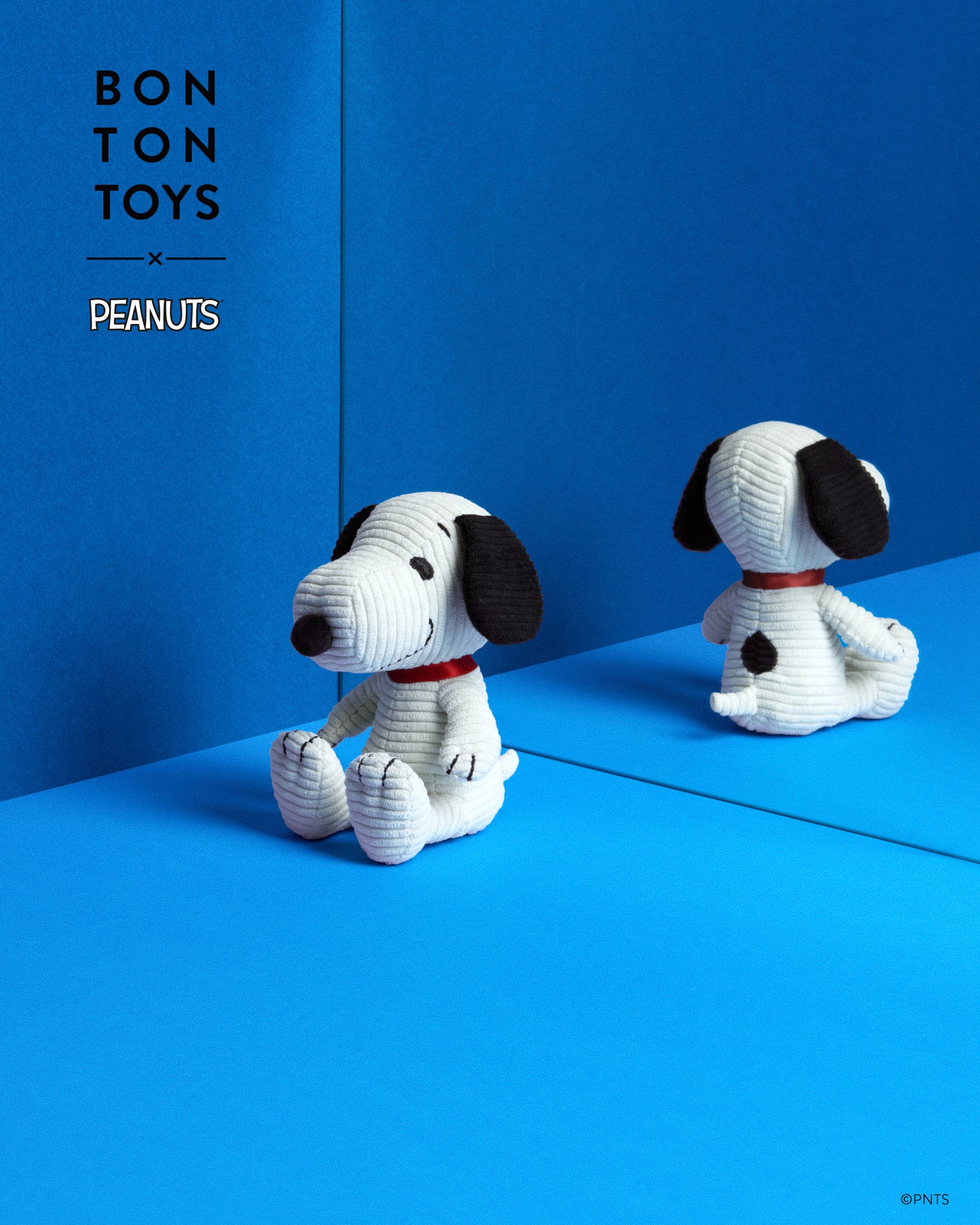 A crocheted plush toy of Snoopy from Bon Ton Toys x Peanuts, shown in a studio setting with its reflection visible in a blue mirror. The background is a solid blue with the 'BON TON TOYS x PEANUTS' logo in the upper left.