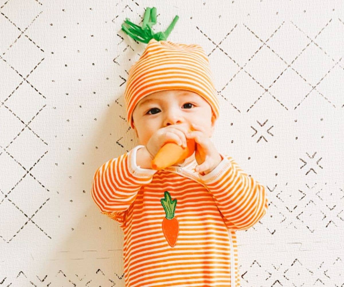 Baby wearing an orange and white striped outfit with a carrot design, holding a carrot toy on a white quilted background.