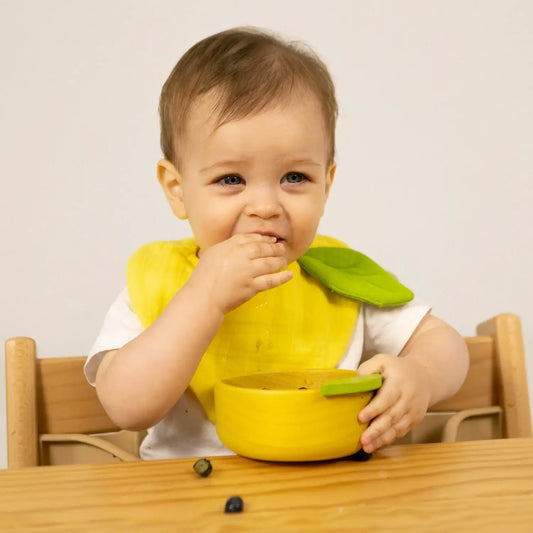 Baby sitting in a high chair with a yellow bib and bowl, eating.